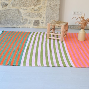 Op de afbeelding: Een rechthoekig vloerkleed met verticale strepen in drie verschillende kleursecties: oranje en groen, lichtroze en groen, en oranje en lichtroze. Het kleed heeft witte franjes aan de korte zijden en een gestructureerd, geweven oppervlak.