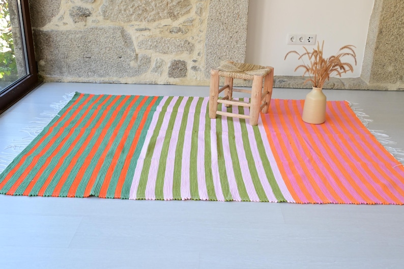 Op de afbeelding: Een rechthoekig vloerkleed met verticale strepen in drie verschillende kleursecties: oranje en groen, lichtroze en groen, en oranje en lichtroze. Het kleed heeft witte franjes aan de korte zijden en een gestructureerd, geweven oppervlak.