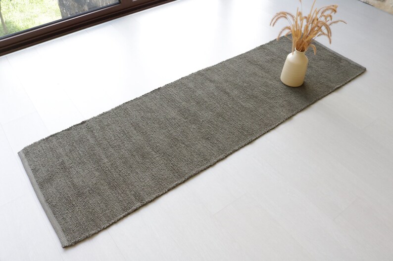 May include: A long, rectangular, gray rug with a textured surface. The rug is on a white floor.