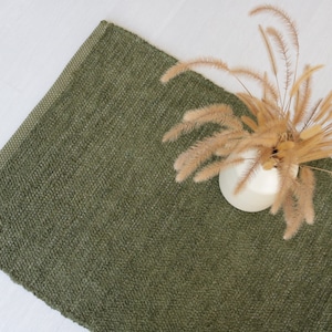 May include: An olive green woven rug with a textured surface. A white ceramic vase holds dried beige grasses, creating a natural, earthy aesthetic. The rug's corner is visible, showcasing its woven pattern.