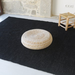 May include: A round, woven, white cushion sits on a black rug. The cushion is made of natural materials and has a slightly textured surface.