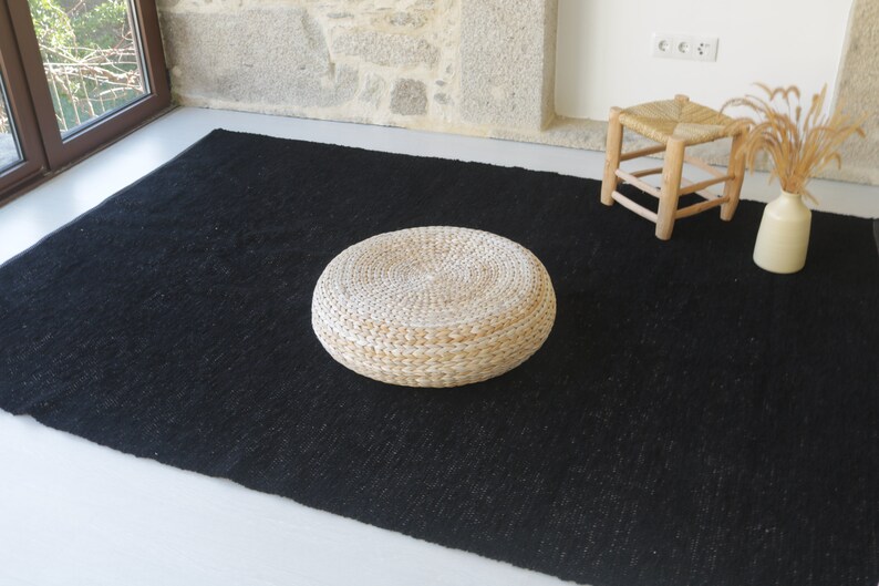 May include: A round, woven, white cushion sits on a black rug. The cushion is made of natural materials and has a slightly textured surface.