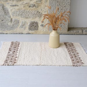 Op de afbeelding: Een rechthoekig, gebroken wit tapijt met bruine patronen en franjes. Een beige vaas met gedroogde planten, geplaatst tegen een stenen muur.