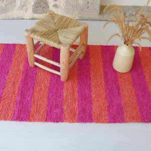 May include: A pink and orange striped rug with a wooden stool and a vase with dried flowers on top.