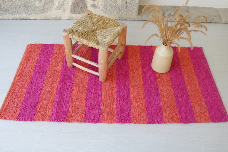 May include: A pink and orange striped rug with a wooden stool and a vase with dried flowers on top.