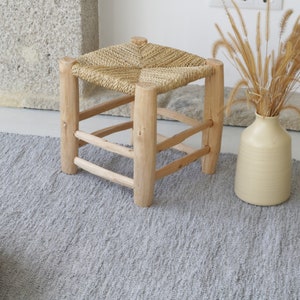 May include: A woven straw seat stool with a wooden frame sits on a gray rug with a woven ottoman in the foreground. A vase with dried grass is in the background.