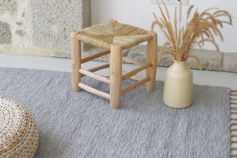 May include: A woven straw seat stool with a wooden frame sits on a gray rug with a woven ottoman in the foreground. A vase with dried grass is in the background.