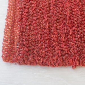 May include: Close-up of a red woven rug with a textured surface. The rug has a fringe edge and is made of a soft, natural material.
