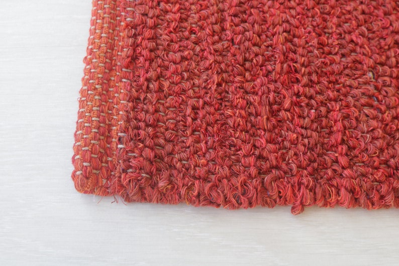 May include: Close-up of a red woven rug with a textured surface. The rug has a fringe edge and is made of a soft, natural material.