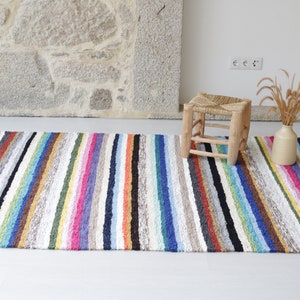 Puede incluir: Una alfombra de rayas coloridas con flecos en los bordes. La alfombra est&aacute; hecha de un material suave y texturizado y tiene una variedad de colores, incluyendo rojo, naranja, amarillo, verde, azul, morado y marr&oacute;n. Un peque&ntilde;o taburete de madera con un asiento tejido se encuentra sobre la alfombra.