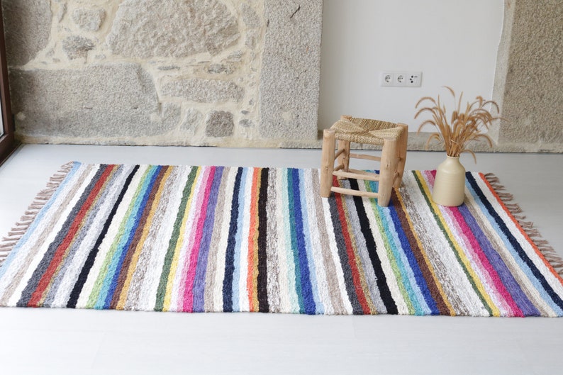 Puede incluir: Una alfombra de rayas coloridas con flecos en los bordes. La alfombra est&aacute; hecha de un material suave y texturizado y tiene una variedad de colores, incluyendo rojo, naranja, amarillo, verde, azul, morado y marr&oacute;n. Un peque&ntilde;o taburete de madera con un asiento tejido se encuentra sobre la alfombra.