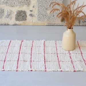 Puede incluir: Una alfombra tejida blanca con rayas rojas. La alfombra tiene un borde con flecos y está sobre un suelo gris claro.