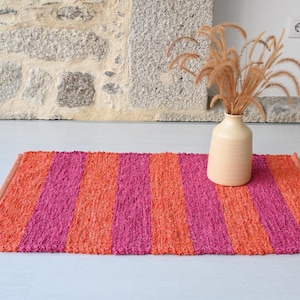 May include: A striped rug in shades of pink and orange, with a beige vase holding dried plants. The rug's design features vertical stripes. The rug is made of a textured material. The vase is ceramic.