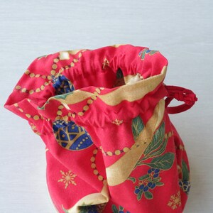 Pode incluir: Uma bolsa de cord&atilde;o vermelha com uma estampa de Natal com ornamentos dourados, folhas verdes e bagas azuis. A bolsa &eacute; feita de tecido e tem um fecho de cord&atilde;o.
