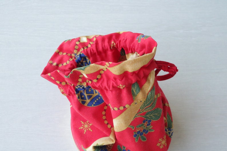 Pode incluir: Uma bolsa de cord&atilde;o vermelha com uma estampa de Natal com ornamentos dourados, folhas verdes e bagas azuis. A bolsa &eacute; feita de tecido e tem um fecho de cord&atilde;o.