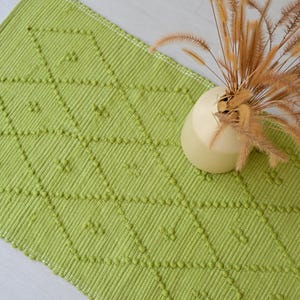 Op de afbeelding: Een groene, getextureerde vloerkleed met een ruitpatroon. Het vloerkleed heeft een herhalend patroon van ruitvormen met kleine, verhoogde details. Een crèmekleurige vaas met gedroogde, bruinachtige grassen staat op het vloerkleed.
