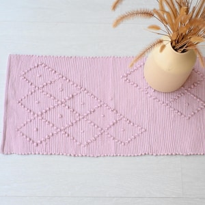 Pode incluir: Um tapete rosa tecido com um padrão de diamante e pompons texturizados. O tapete está sobre um piso de madeira branca.