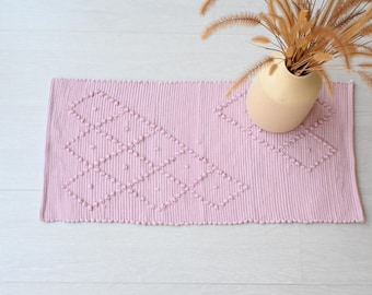 Può includere: Un tappeto rosa tessuto con un motivo a rombi e pompon texturizzati. Il tappeto è su un pavimento in legno bianco.