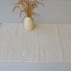 Puede incluir: Una alfombra tejida blanca con flecos en todos los lados. La alfombra est&aacute; tendida sobre una superficie gris claro. Un jarr&oacute;n con hierba seca est&aacute; sentado encima de la alfombra.