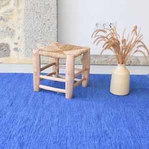 May include: A blue woven rug with a wooden stool with a woven seat. The stool is in the center of the rug. A vase with dried grass is on the right side of the rug.