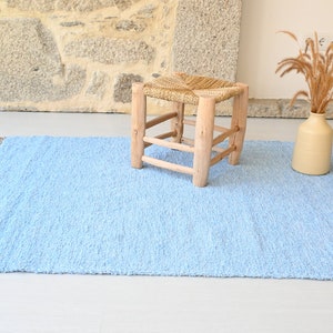Pu&ograve; includere: Un tappeto blu chiaro con frange sui bordi. Il tappeto &egrave; realizzato in un materiale morbido e testurizzato e ha una lucentezza sottile. Uno sgabello in legno con una seduta intrecciata si trova sul tappeto.