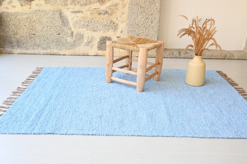 Pu&ograve; includere: Un tappeto blu chiaro con frange sui bordi. Il tappeto &egrave; realizzato in un materiale morbido e testurizzato e ha una lucentezza sottile. Uno sgabello in legno con una seduta intrecciata si trova sul tappeto.
