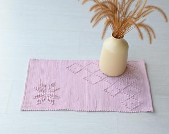 Può includere: Un tappeto rosa intrecciato con un motivo a rombi e un design a stella. Un vaso in ceramica beige con erba secca è sopra il tappeto.