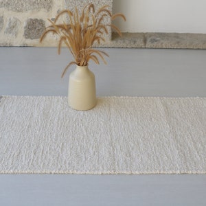 Puede incluir: Una alfombra blanca y esponjosa con flecos en los bordes. La alfombra est&aacute; colocada sobre un suelo gris claro con un jarr&oacute;n de hierba seca en el fondo.