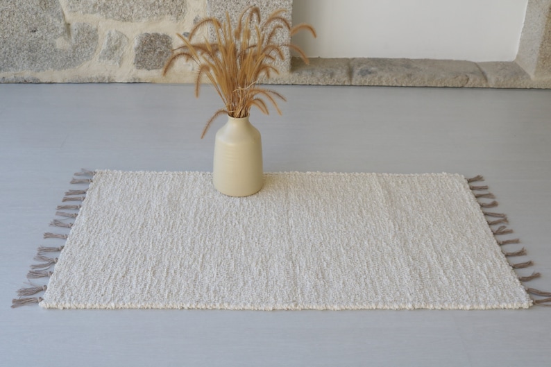 Puede incluir: Una alfombra blanca y esponjosa con flecos en los bordes. La alfombra est&aacute; colocada sobre un suelo gris claro con un jarr&oacute;n de hierba seca en el fondo.