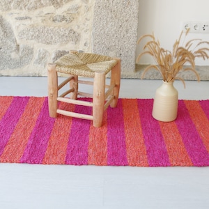 May include: A pink and orange striped rug with a woven texture. A small wooden stool with a woven straw seat sits on the rug.