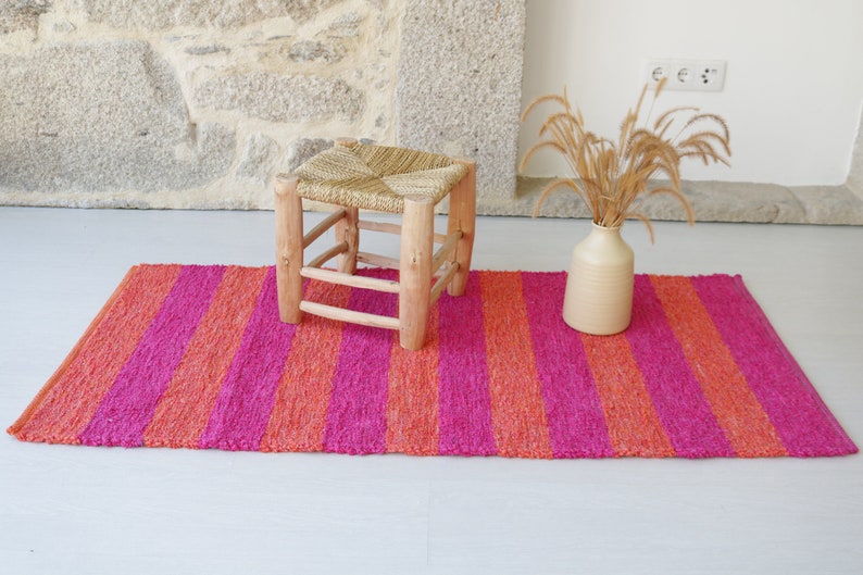 May include: A pink and orange striped rug with a woven texture. A small wooden stool with a woven straw seat sits on the rug.