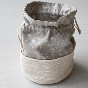 Pu&ograve; includere: Un cestino rotondo in tessuto bianco sporco con un sacchetto grigio con coulisse floreale all'interno. La borsa presenta un motivo botanico bianco. Il cestino &egrave; realizzato in materiale spesso e tessuto e la borsa ha una chiusura con coulisse.