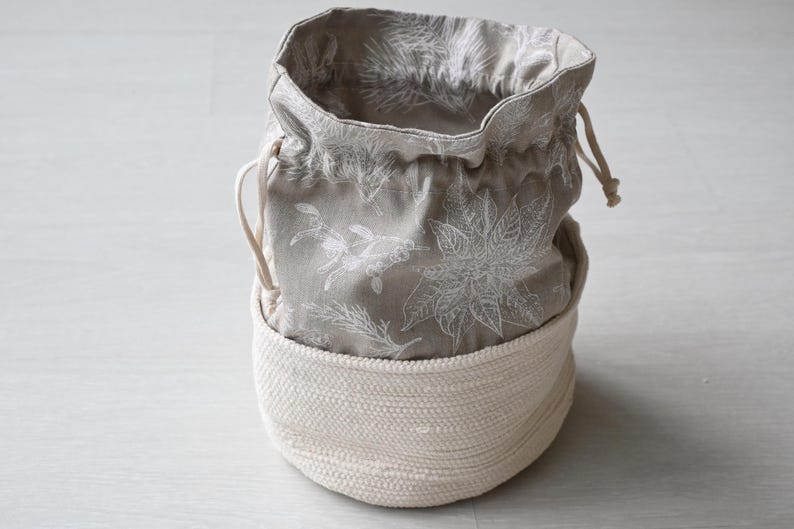 Pu&ograve; includere: Un cestino rotondo in tessuto bianco sporco con un sacchetto grigio con coulisse floreale all'interno. La borsa presenta un motivo botanico bianco. Il cestino &egrave; realizzato in materiale spesso e tessuto e la borsa ha una chiusura con coulisse.