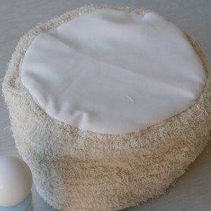 May include: A round, cream-colored pouf with a white fabric top. The pouf is made of a fluffy, textured material.