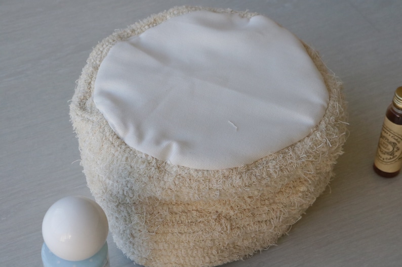 May include: A round, cream-colored pouf with a white fabric top. The pouf is made of a fluffy, textured material.