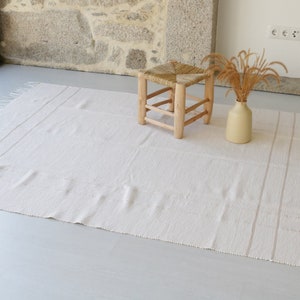 Puede incluir: Una alfombra blanca tejida con flecos en los bordes y rayas sutiles. La alfombra est&aacute; colocada sobre un suelo gris claro con un taburete de madera y un jarr&oacute;n con hierba seca.