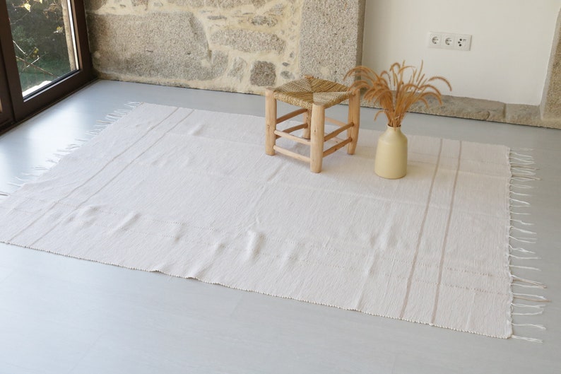 Puede incluir: Una alfombra blanca tejida con flecos en los bordes y rayas sutiles. La alfombra est&aacute; colocada sobre un suelo gris claro con un taburete de madera y un jarr&oacute;n con hierba seca.