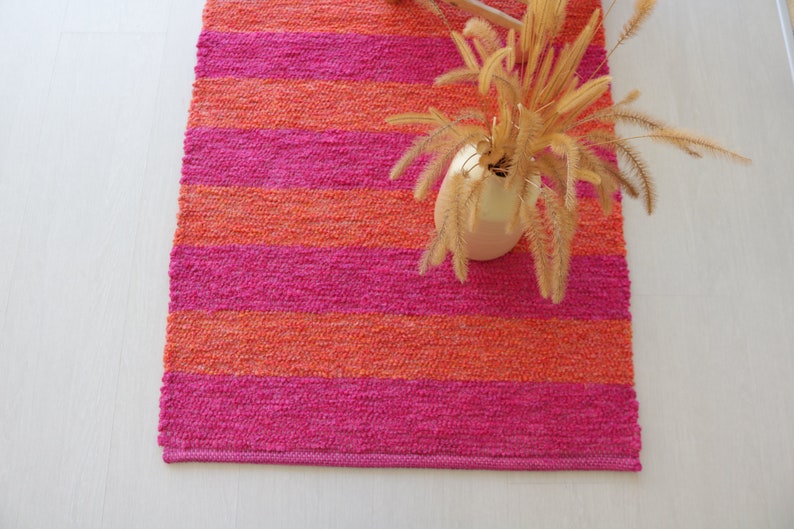 May include: A pink and orange striped woven rug with a vase of dried flowers on top.