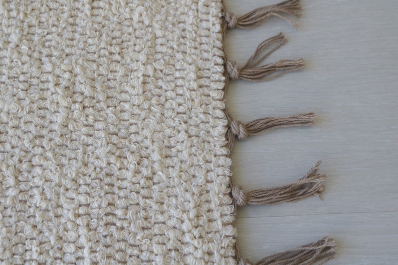 Puede incluir: Una alfombra tejida de color crema con un borde de flecos. Los flecos est&aacute;n hechos de una fibra natural, como el yute o el c&aacute;&ntilde;amo, y son de color marr&oacute;n claro.