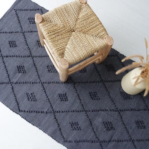May include: A dark gray woven rug with a diamond pattern and a woven wood stool with a straw seat.