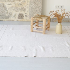 Puede incluir: Una alfombra blanca tejida con flecos en los bordes y un taburete de madera natural con asiento tejido.