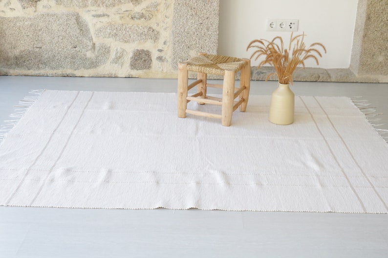 Puede incluir: Una alfombra blanca tejida con flecos en los bordes y un taburete de madera natural con asiento tejido.