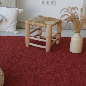 May include: A red woven rug with a woven stool and a vase with dried grass on top.
