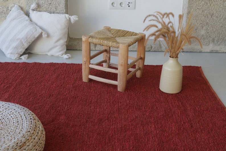 May include: A red woven rug with a woven stool and a vase with dried grass on top.