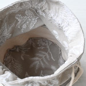 Pu&ograve; includere: Una borsa beige con coulisse con un motivo floreale bianco all'esterno e all'interno. L'interno della borsa &egrave; di colore beige chiaro. La borsa &egrave; aperta, rivelando l'interno, e il cordoncino &egrave; visibile.