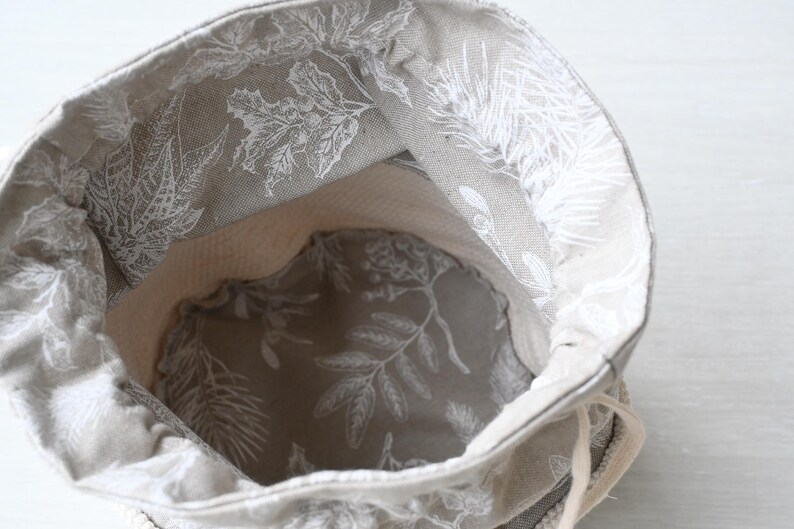 Pu&ograve; includere: Una borsa beige con coulisse con un motivo floreale bianco all'esterno e all'interno. L'interno della borsa &egrave; di colore beige chiaro. La borsa &egrave; aperta, rivelando l'interno, e il cordoncino &egrave; visibile.