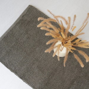 May include: A gray, textured rug with a vase of dried pampas grass on top. The rug is rectangular and has a fringed edge.