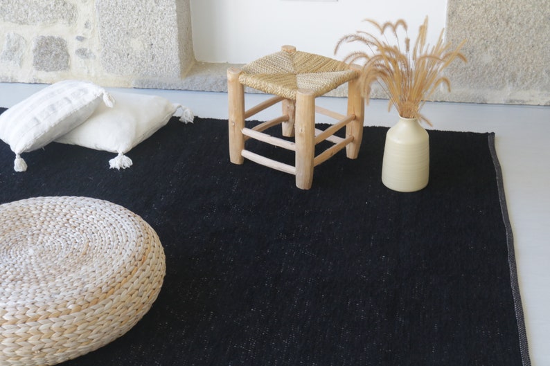 May include: A black woven rug with a white woven stool, a white woven pouf, and a white vase with dried grass.