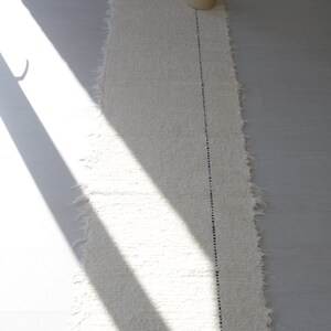 May include: A white woven rug with a black line down the center. The rug has a frayed edge and is laying on a white floor.