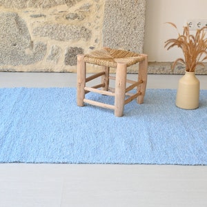 Pu&ograve; includere: Un tappeto blu chiaro con frange sui bordi. Uno sgabello di legno con una seduta intrecciata si trova sul tappeto. Un vaso con erba secca si trova sul tappeto.